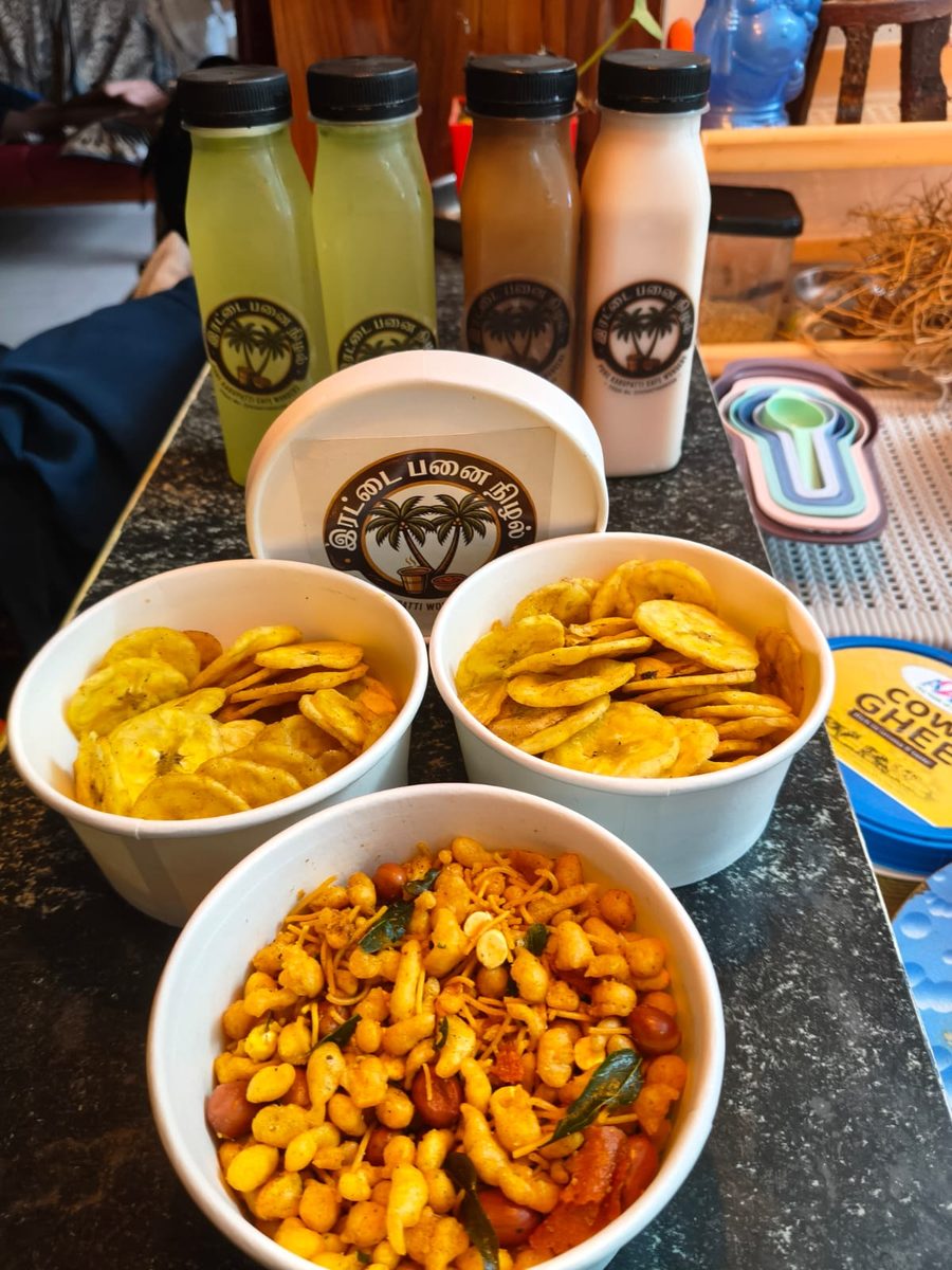Irattai Panai Nizhal branded product spread — lime juice, karupatti sarbath, rose milk bottles with banana chips and sundal snack bowls in branded containers